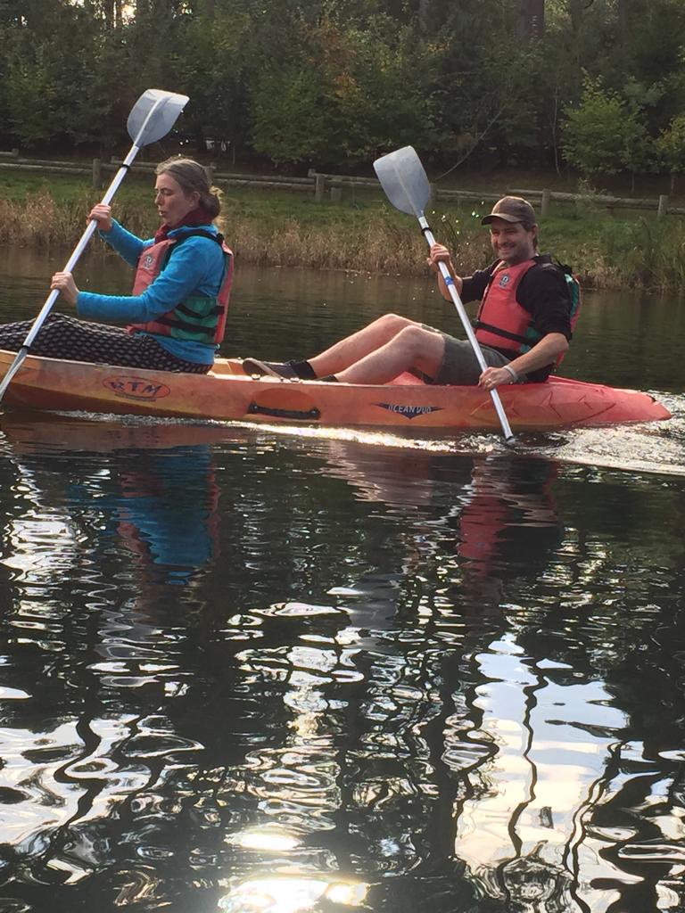 A photo of me and my husband in a kayak on a lake, he's smiling at the camera and I'm concentrating on my paddle