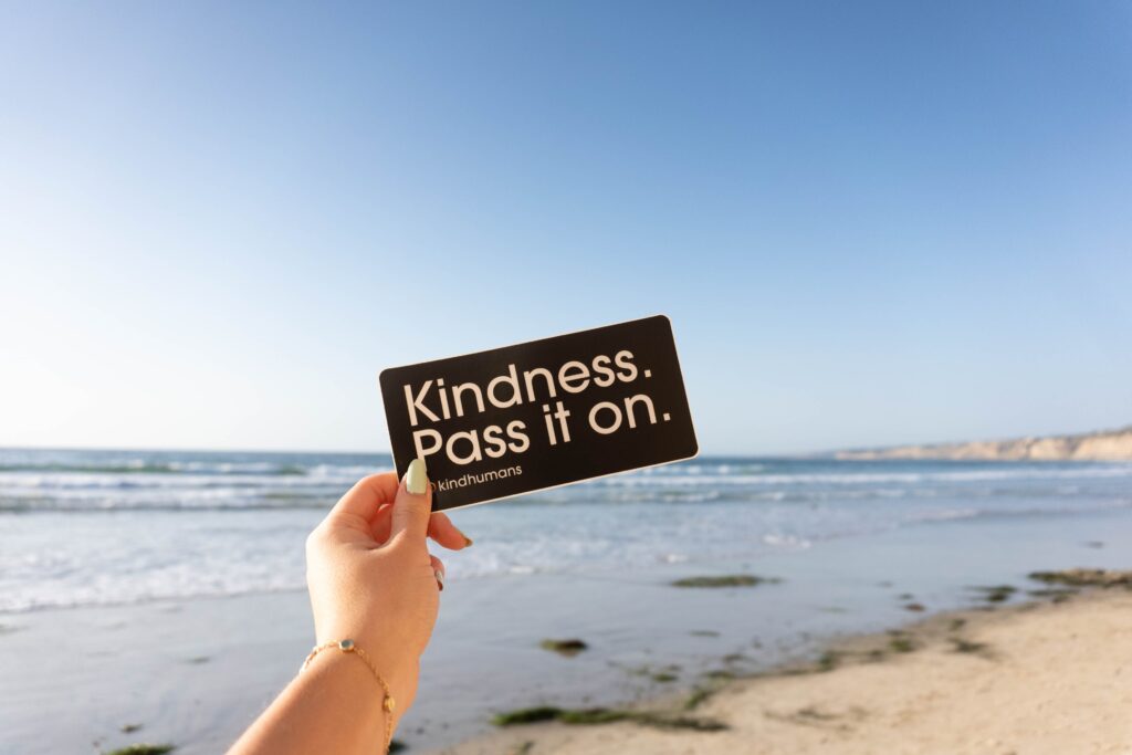 A hand at the beach holding out a sign that says 'Kindness. Pass it on'