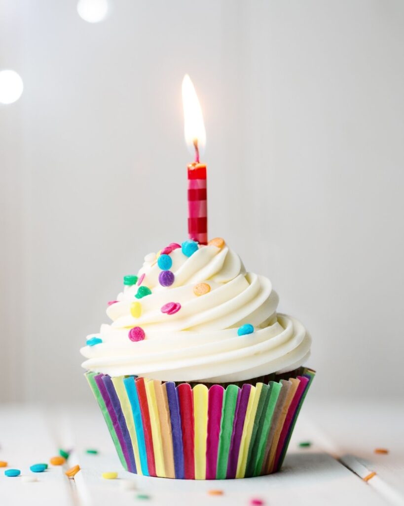 A white cupcake in a rainbow case with white icing and a single candle lit in the middle of it.