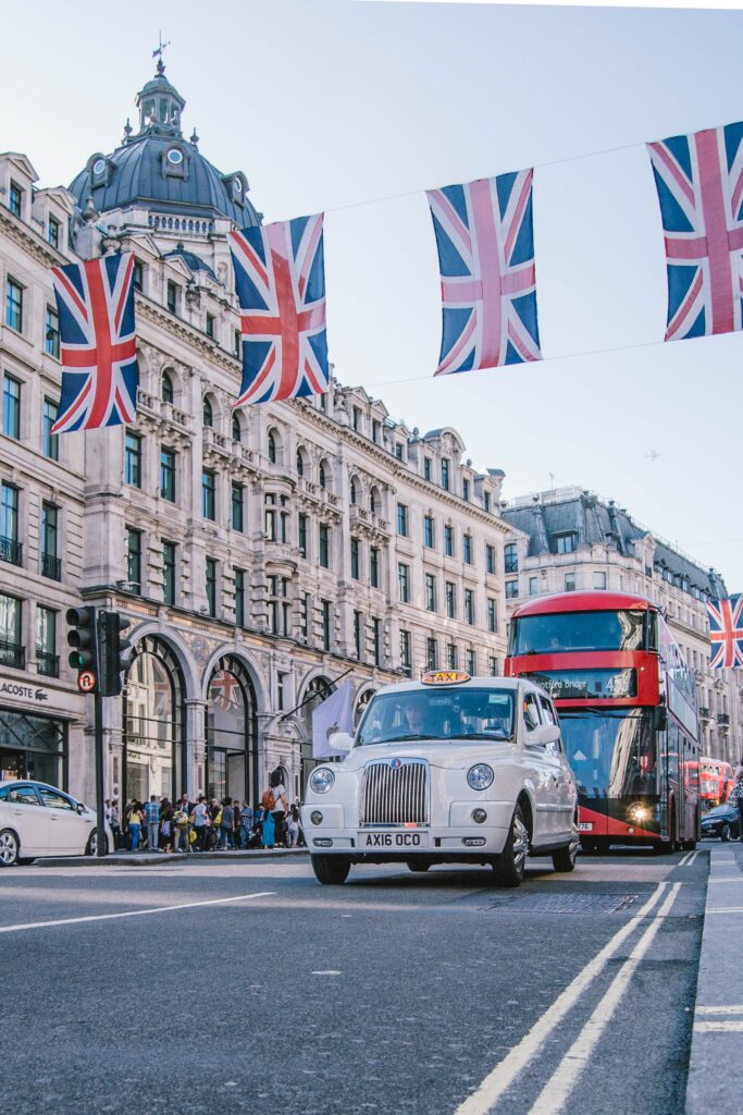 a white London taxi drives along a street in front of a red double decker bus. Union Jack flags are strung across the street.
