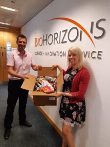 Tris and Naomi hold a large cardboard box filled with crisp packets between them in front of a wall with the BioHorizons logo on it.]