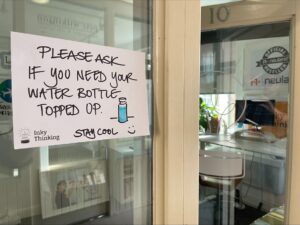 A hand written sign on an office door which reads “please ask if you need your water bottle topped up. Stay cool.” A bottle of water and a smiley face have been drawn on the sign.