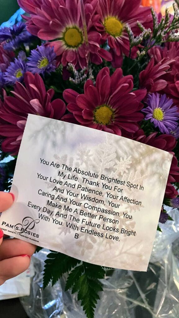 a bunch of red, purple and other deep coloured flowers in full bloom behind a note with the words in the caption on it.