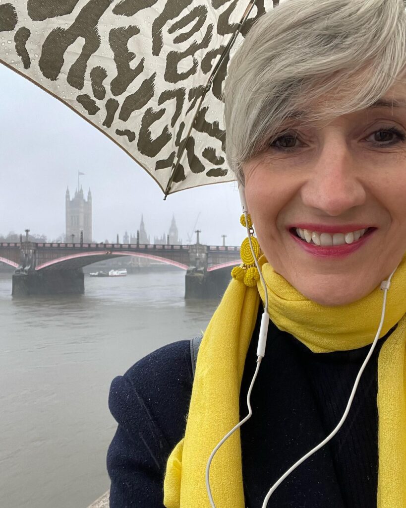 a femme presenting person with short blonde hair swept across their face and a yellow scarf wrapped around their neck, standing in front of the Thames on a rainy day with an umbrella up.