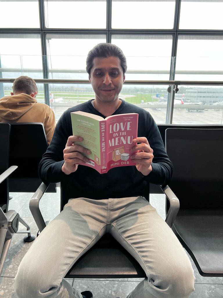 Mimi's husband, a man with medium light skin tone sits on an airport chair, planes can be seen in the background through the window, reading Love On The Menu by Mimi Deb.