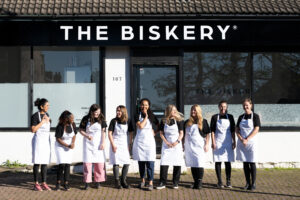 Nine women wearing white aprons and black tshirts, stand outside The Biskery, laughing and looking at each other.