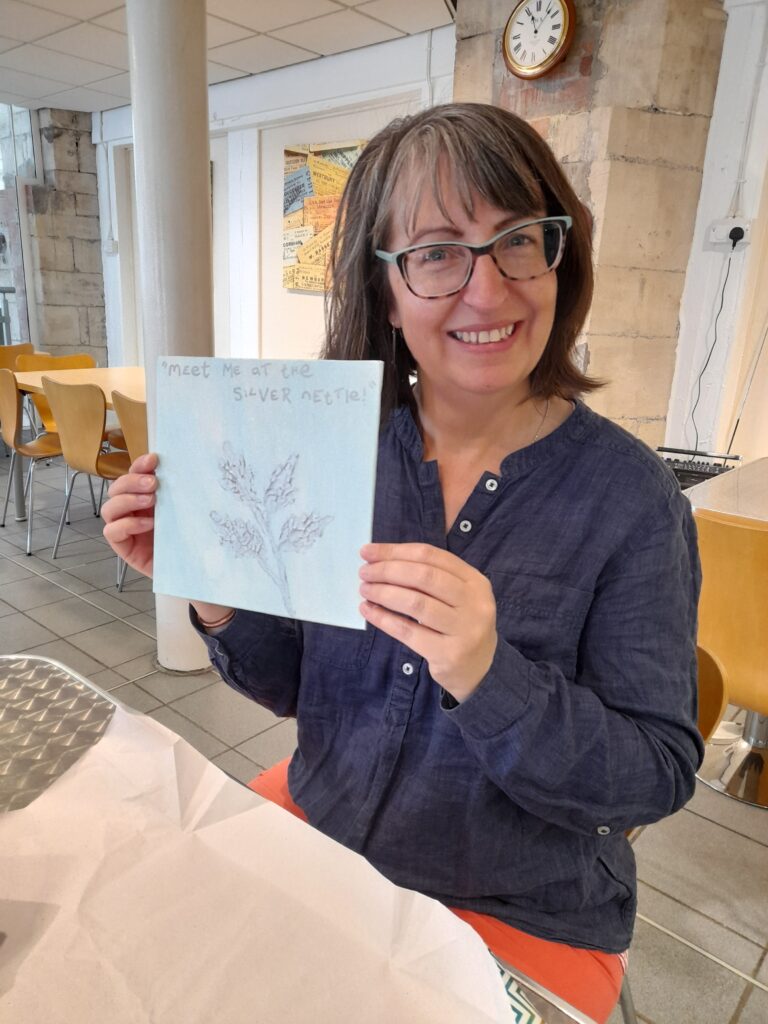 Morwhenna, a woman with light skin tone and shoulder length dark hair, wearing glasses. She is sat down at a table and holding up a drawing of a plant with writing above it that says "meet me at the silver nettle."