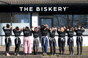 A row of 9 people standing with their backs to the camera. They are all wearing black tops with the words Kindness in Biscuit Form printed on the back and are pointing over the shoulders to the words. They stand in front of The Biskery bakery building.