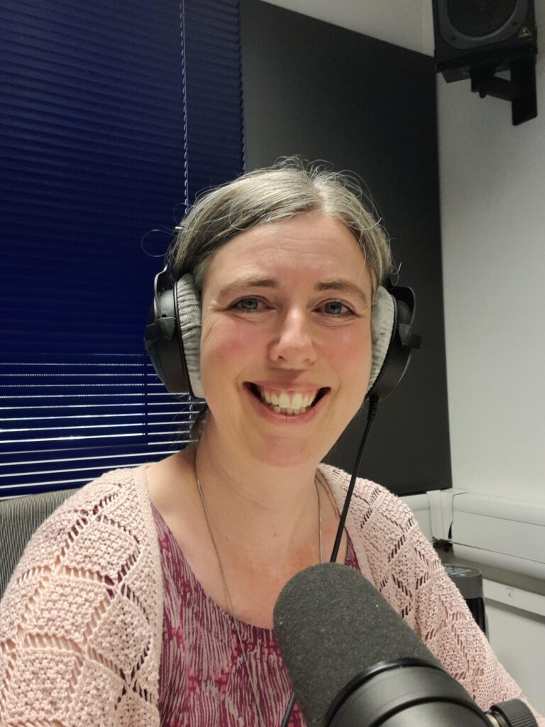 Sarah smiling broadly wearing headphones with a large mic in front of her.