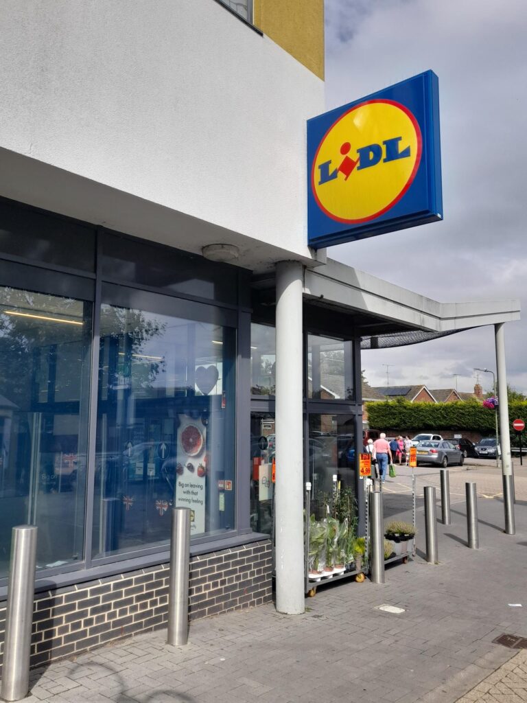 A Lidl show viewed from outside. The car park is visible beyond the doors with people walking to and from their cars.