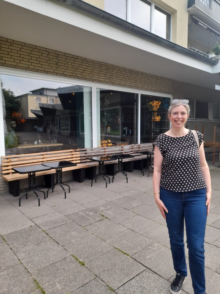 Sarah standing outside a cafe on a paved street. There are wooden benches alongside the window of the cafe with small tables in front of them.