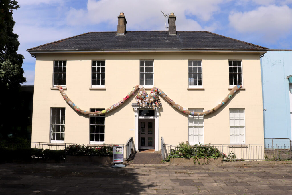 The Flowerfield Arts Centre, a white building with lots of windows. The Woven hug is a pair of textile arms which look as thought they are coming out of first floor windows on either side of the building and hugging it, with the hands joining over the front door.