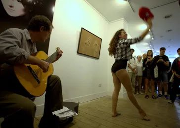 an art gallery with the artist, Lawrence, sitting on the left of the photo playing a guitar. He is wearing dark trousers and a grey shirt. On the right is a female dancer in a black leotard and a black and white check shirt and long dark hair dancing in front of a painting. There are people standing around watching her.