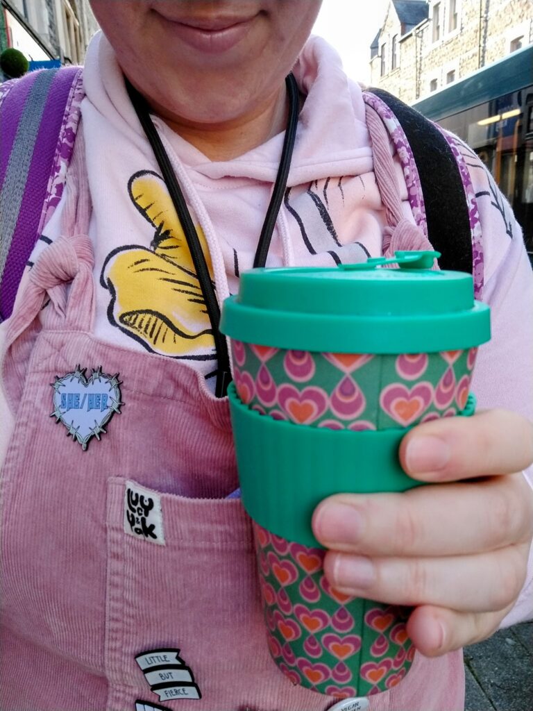 a person in a pair of pink Lucy and Yak cord dungarees with a pink Dumbo Disney hoodie under them, carries a green reusable coffee cup with pink hearts on it. On the dungarees are some pin badges, one reading "she/her," the other reading "little but fierce."