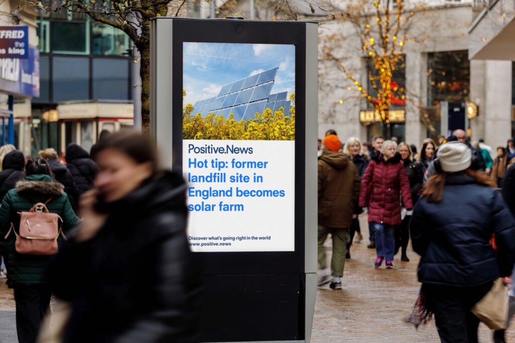 An outdoor screen on a busy town centre showing a Positive News headline reading “hot tip: former landfill site in England becomes solar farm.”