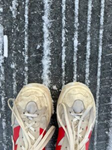 a pair of trainers on someone’s feet standing on a tarmac road with lines of snow on it.