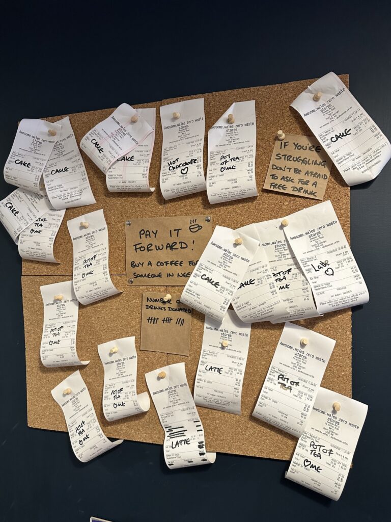A cork board on a blue wall with 21 pay it forward receipts on it and some hand written notes reading “if you’re struggling, don’t be afraid to ask for a free drink. Pay it forward, buy a coffee for someone in need.” And a tally chart of pay it forward drinks.