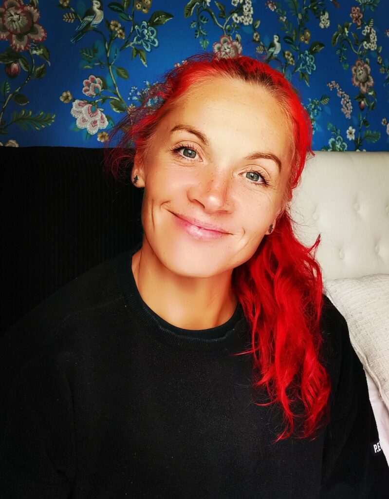 a woman with bright red hair tied back but stretching over her shoulder, wearing a black jumper. She is sat on a chair and smiling having received a random act of kindness.