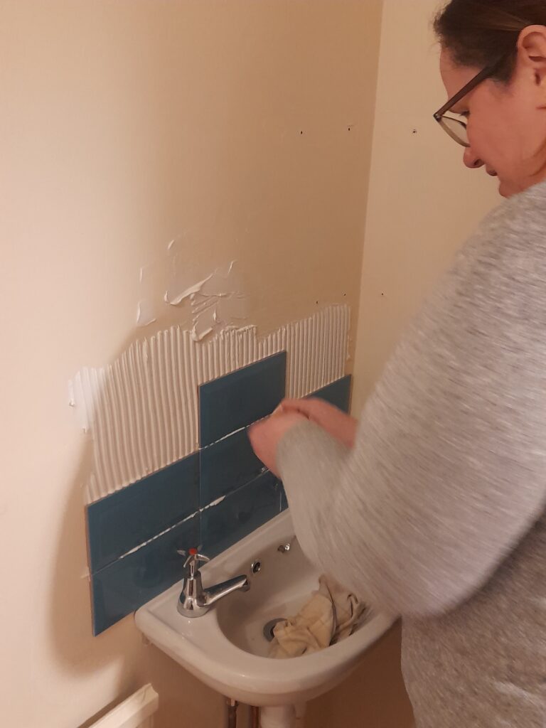 Di, a woman with light skin tone wearing glasses, tiling a small area above a sink. Two rows of blue tiles and one on the third row are in place with some white grout all around ready to have more tiles applied.