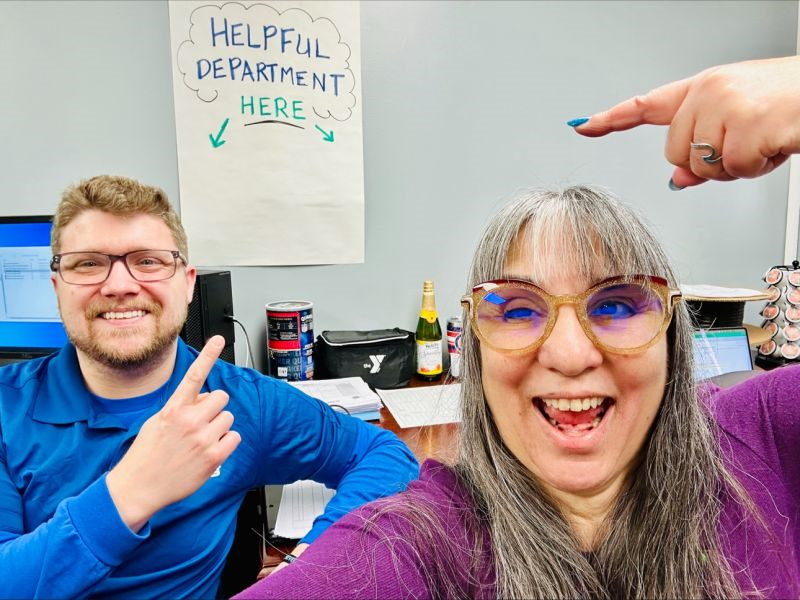 Karyn takes a selfie with Joshua behind her. Karyn is a woman with long grey hair wearing a purple top and round glasses with yellow rims. She smiles broadly and points to a sign which reads “helpful department here” behind her. Joshua is a man with short strawberry blonde hair and a goatee wearing a blue top and black rimmed glasses. He is smiling broadly and pointing to the same sign.