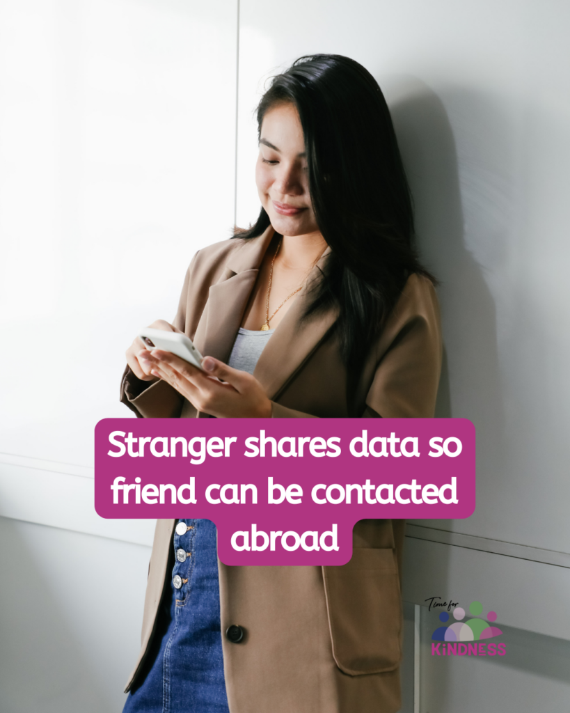 a woman with long black hair, wearing a brown coat and denim skirt. She is holding a phone and smiling while leaning against a white wall. Text overlaid reads "Stranger shares data so friend can be contacted abroad.”