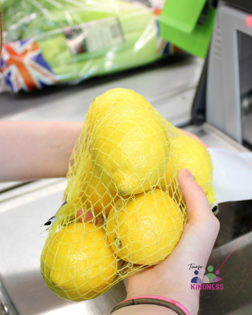 A pair of hands holds a net of lemons to scan them at a supermarket. Beyond the hands is a conveyor belt of shopping.