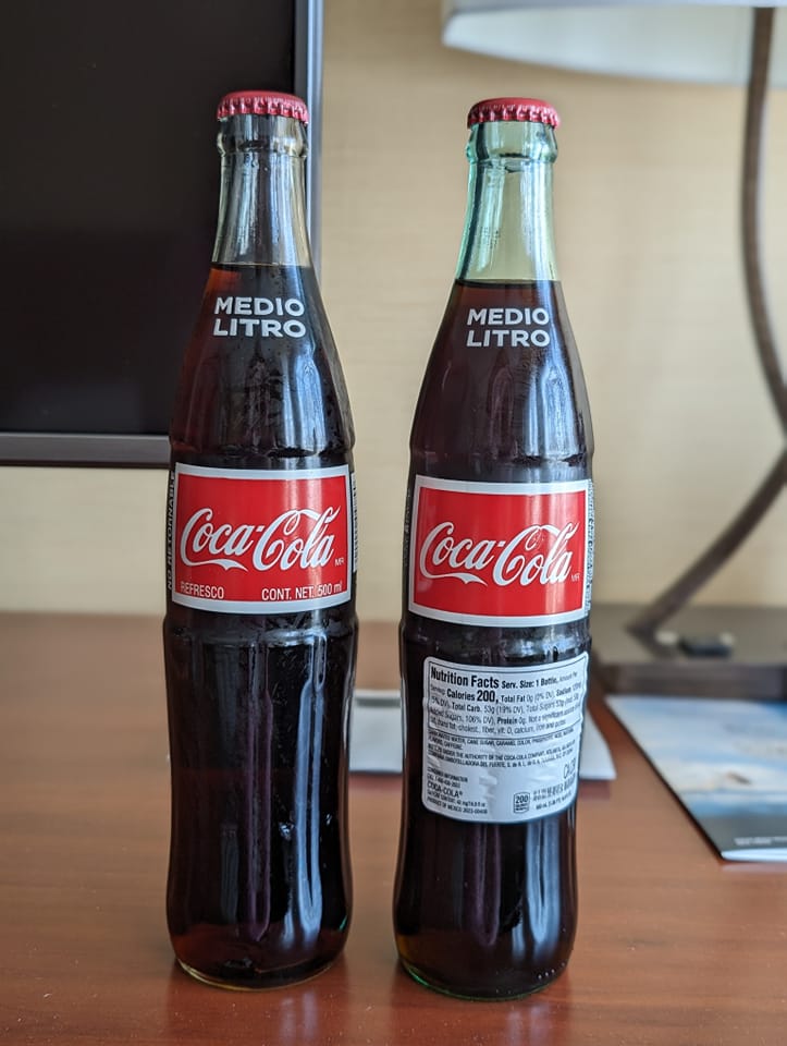 Two almost identical glass bottles of Coca Cola on a wooden table. The glass on the left is clear whereas on the right there is a slight green tinge, the bottle on the right also has an extra information sticker on it. The Coca-Cola label on the left bottle has two extra bits of information under the logo which are not on the bottle on the right.