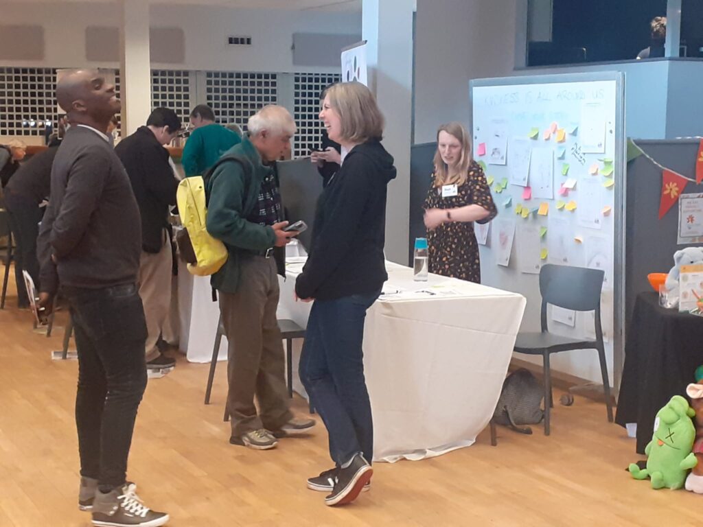 Sarah talking to a man with dark skin tone at Reading Community Festival. Behind them are people milling around the Time for Kindness stand and a large whiteboard with post it notes and drawings all over it.