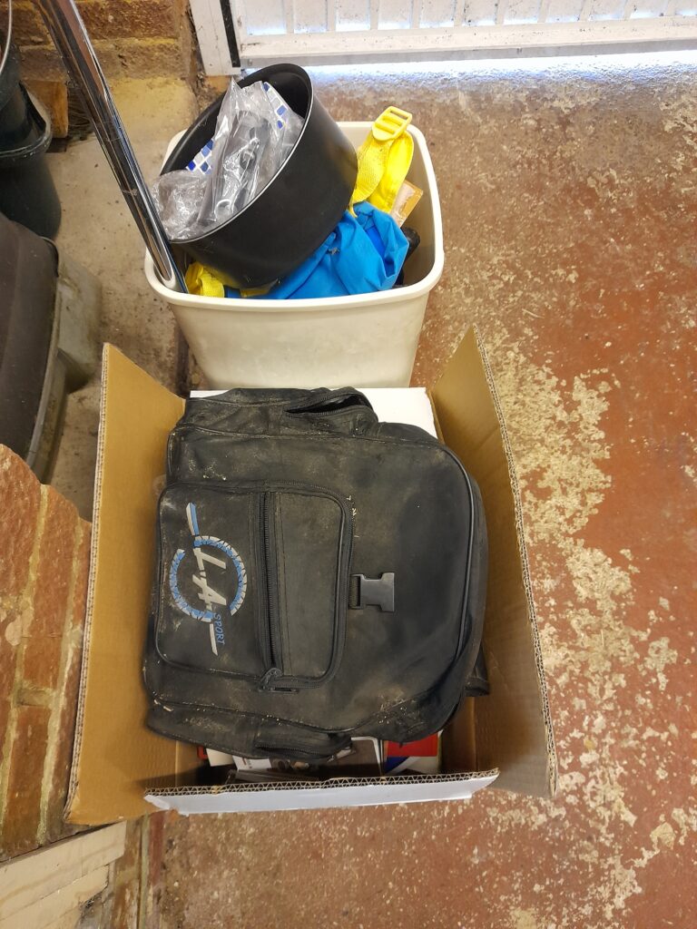 Two boxes filled with things sitting on a floor. In the cardboard box a dusty bag sits on top of some other unidentifiable items ready for the tip. In the plastic box are some rubber gloves, a metal pole and other items.