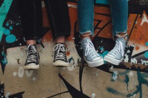 2 pairs of legs and feet belonging to young people, shown wearing trainers and hanging down in front of a wall with graffiti on it