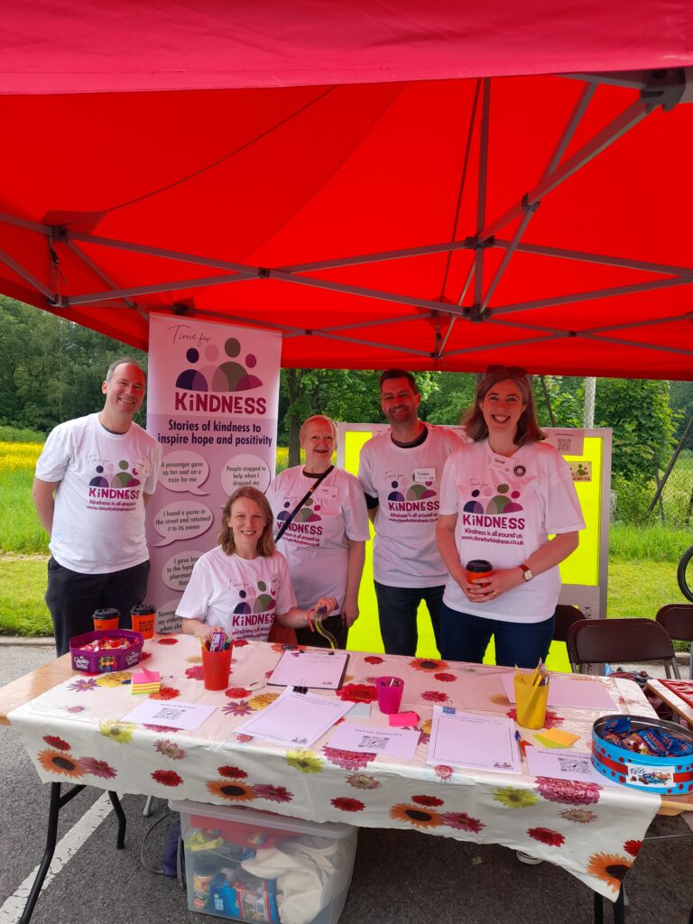 5 members of team Time for Kindness standing under a red gazebo behind a table covered in post its, papers and pens.