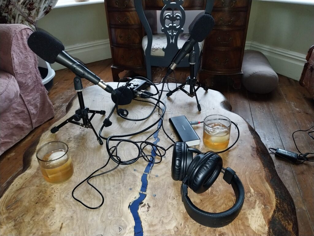 A wooden coffee table set up for a curious chats and kind acts episode with two microphones, two glasses of something with ice in it, headphones, and several wires. with two microphones, two glasses of something with ice in it, headphones, and several wires.
