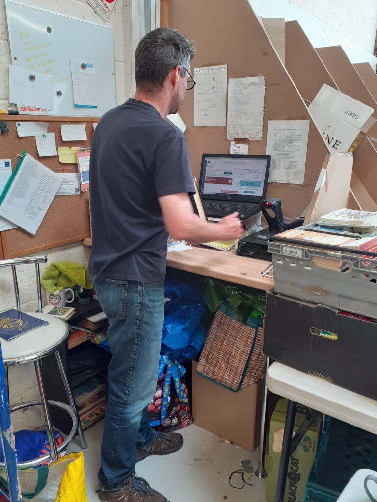 Tris, a man with short dark hair stands with his back to the camera and a computer on a desk in front of him scanning in books. To the side of him is a crate full of books.