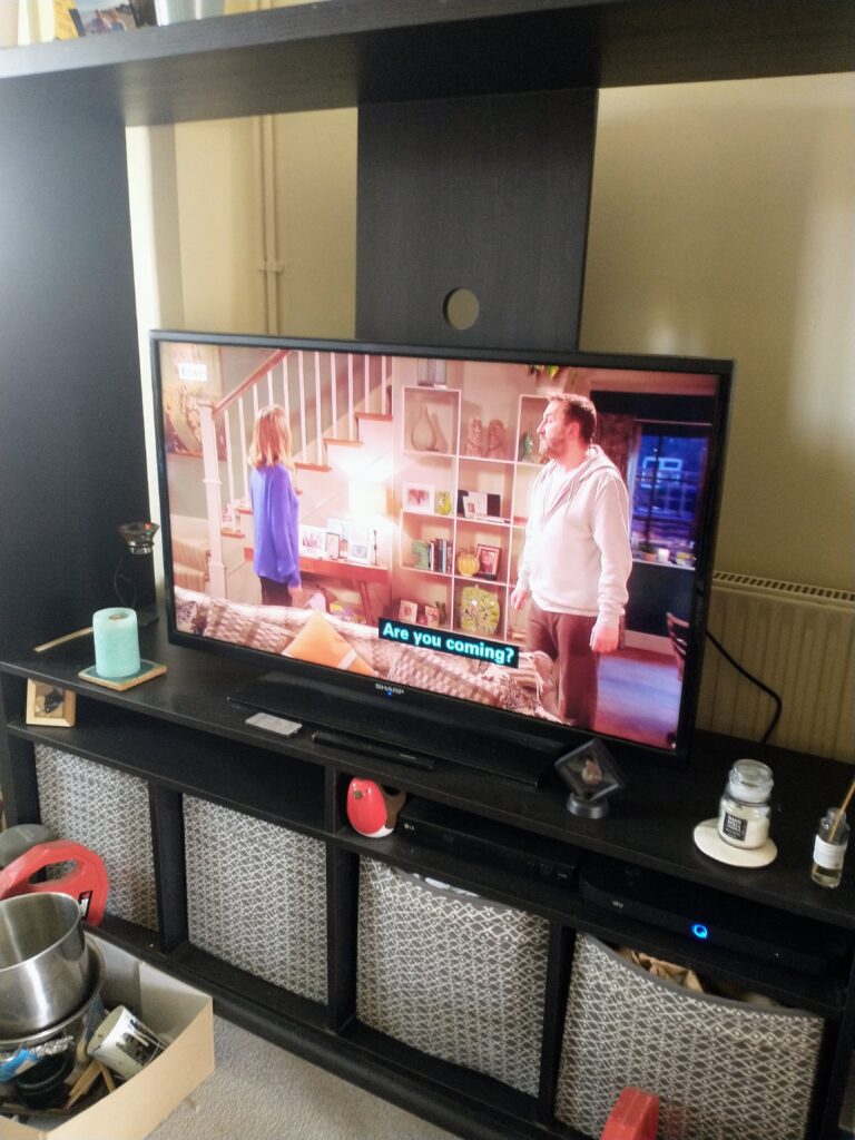 A TV playing a Not Going Out with Lee Mack, there are subtitles on the screen.