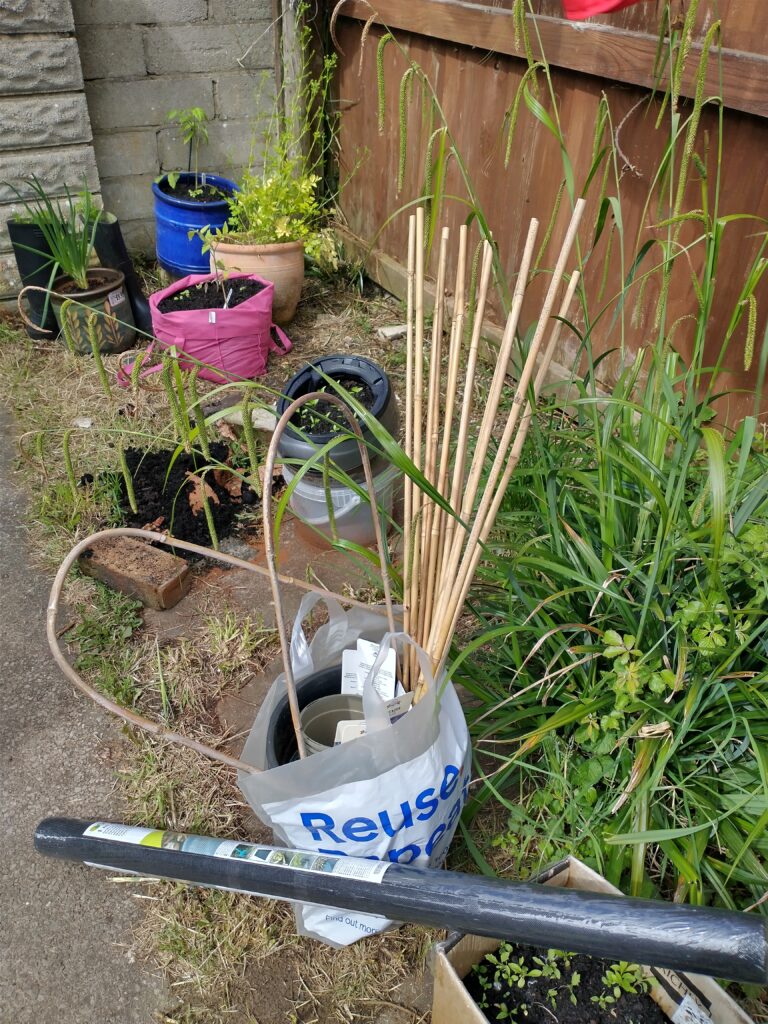 a plastic bag filled with bamboo canes, two bamboo U shaped canes, large and medium sized plastic plant pots. A roll of black garden mesh leaning on a cardboard box of plants. Behind the bag are different containers with plants growing in them.