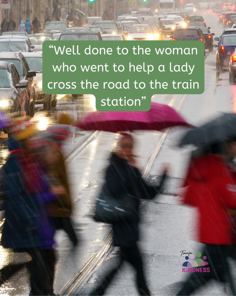 a busy road in rainy weather with cars backed up and blurry people crossing the road, many with umbrellas up. Text overlaid reads “Well done to the woman who went to help a lady cross the road to the train station.”