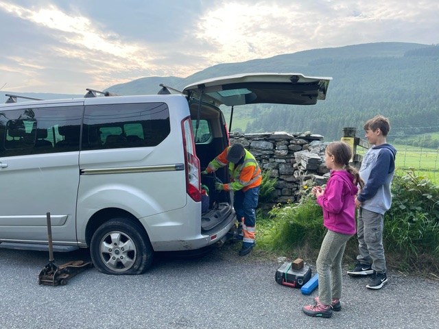 a silver minivan on the side of a rural North Wales road with a Greenflag man working in the boot of it as two children look on.