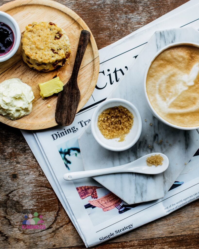 A wooden table with a newspaper on it. On the newspaper is a coffee, a small bowl of sugar and a spoon, and a wooden plate with a scone, butter, jam and cream on it.