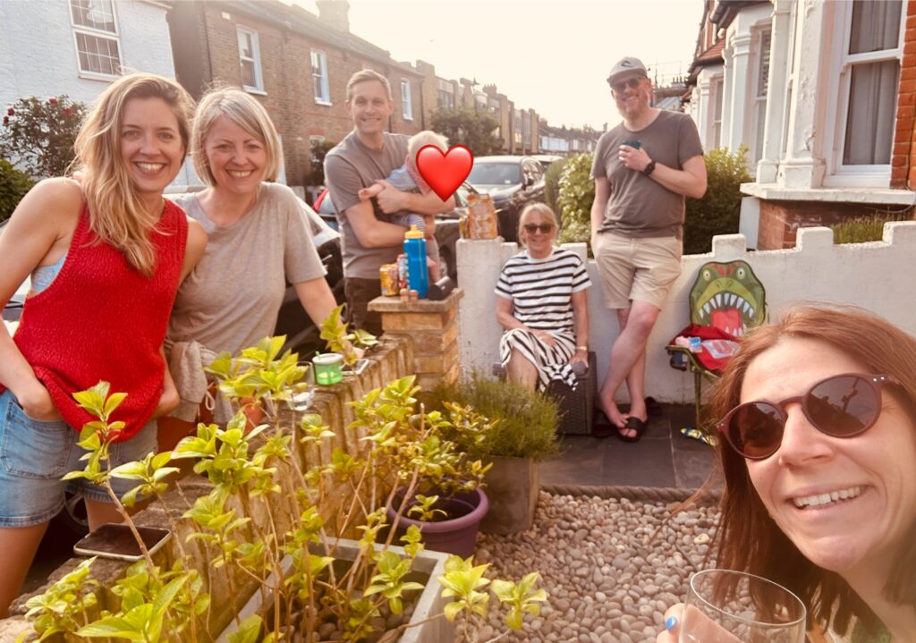 7 people of varying ages, all with light skin tone, sitting or standing in a small paved courtyard on a residential street, some are standing the other side of the wall on the pavement. All are smiling, some holding glasses, one holding a baby.