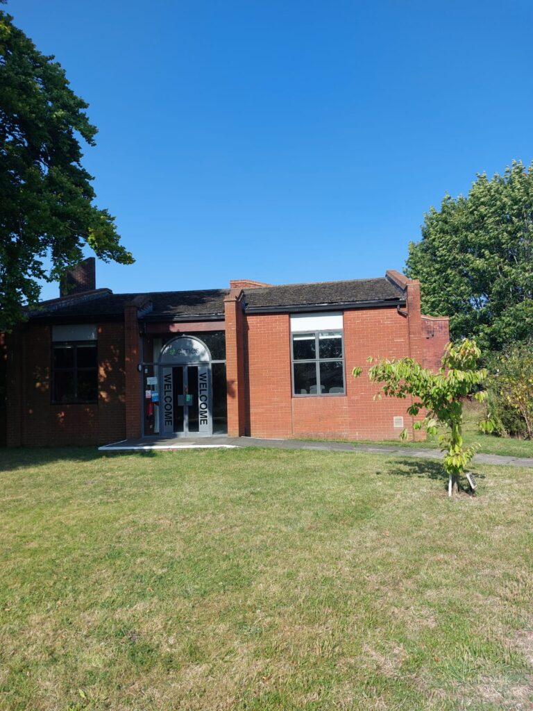 A red brick building with large white door in the shade of a tree with the word “welcome” down the side of it on a sunny day with a totally blue sky above it.