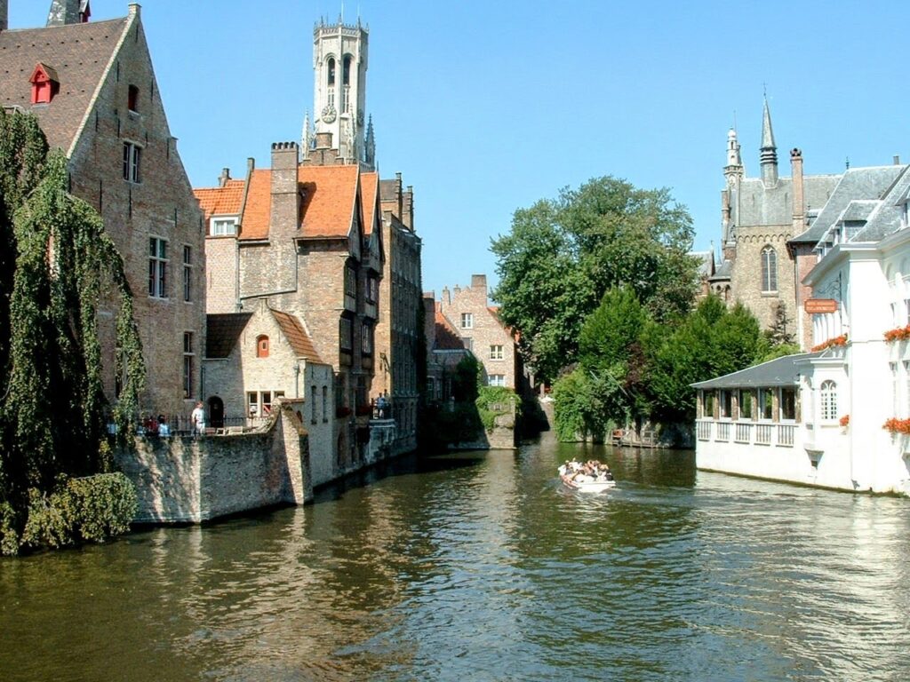 A canal in Brussels with a small white boat on it filled with people. Either side of the canal are buildings. The sky is blue and cloudless