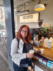 Fran, a woman with light skin tone and long auburn hair, stands in front of FCB coffee holding two take out cups. Behind her is professional coffee making paraphernalia.