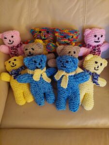 10 small knitted teddies sitting on a chair, there are two of each colour way; yellow, blue, brown, pink, and rainbow.