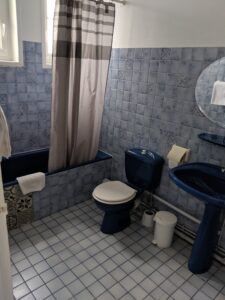 a bathroom; the walls are tiled with small blue tiles and the floor with small white tiles. The basin, toilet and bath are dark blue. There is a mirror above the basin and a grey shower curtain hanging in the bath.