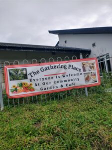 A short metal fence with a large sign on it which says “The Gathering Place. Everyone is welcome at our community garden and cafe.”