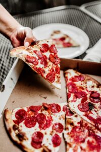 a pepperoni pizza in a box with the hand of someone taking a slice. Just visible outside the box is a plate with a slice of pizza on it