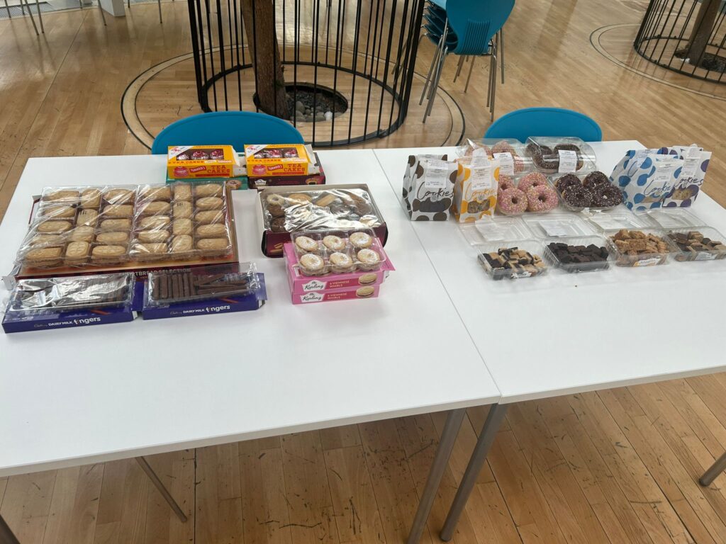 Two tables pushed together with a variety of snacks on them including doughnuts, packs of biscuits, cookies, bite size flapjacks and caramel chocolates, tea cakes and other things.