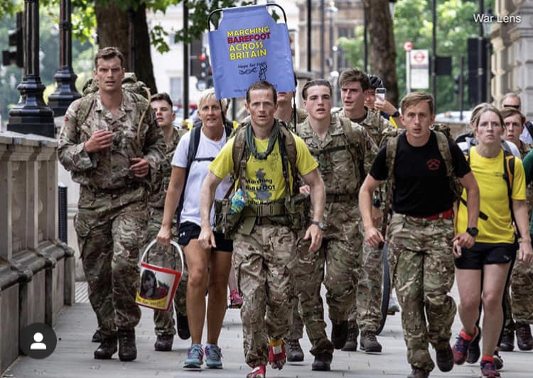 A group of about 15 people, many in army uniforms, one front and centre is not wearing shoes. They are all walking towards the camera down a wide paved area with a short wall to the side. One of them is holding a sign which reads “marching barefoot across britain.”