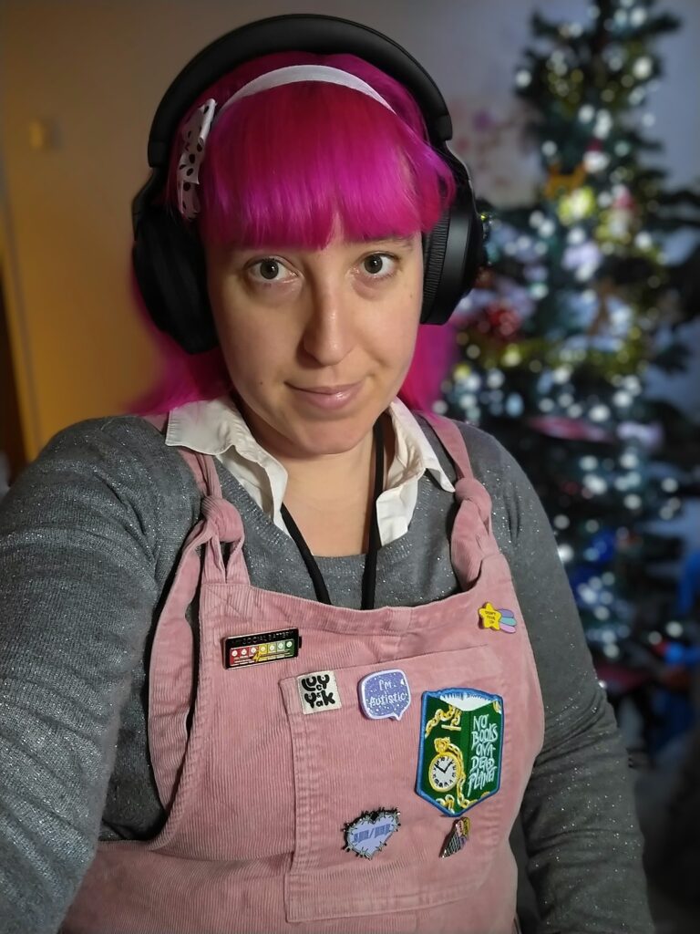 Alexis, a woman with light skin tone and bright pink hair with a white ribbon keeping it back but her fringe forward. She is wearing black headphones, a white shirt with a glittery silver jumper over it, and pink dungarees. Behind her is a Christmas tree.