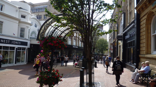 Bournemouth town centre on a sunny day. Some people are sat on a bench to the right. There are some trees and plants and a glass archway in the centre.